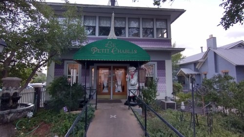 A charming purple house with a green awning reading "Le Petit Chablis," surrounded by greenery and decorative elements.
