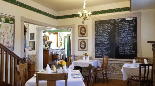 Cozy restaurant interior with white tablecloths, wooden chairs, and a chalkboard menu on the wall. Flowers on tables.