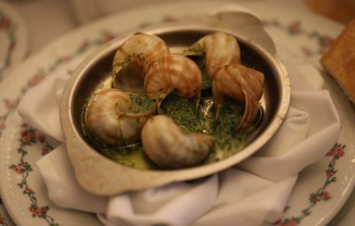 A dish of escargot in garlic butter, served in a metal dish on a floral-patterned plate with a napkin.