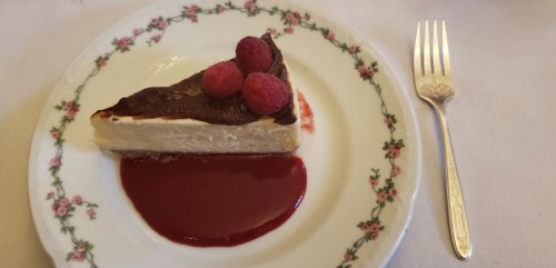 A slice of cheesecake topped with chocolate and raspberries, served on a floral-patterned plate with raspberry sauce.