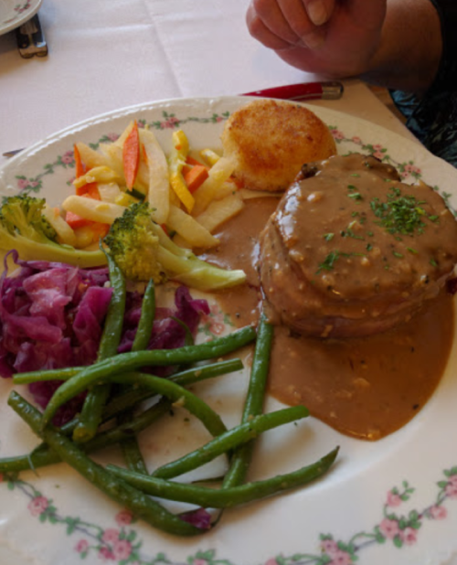 A plate with a meat dish topped with gravy, green beans, broccoli, red cabbage, and apple slices, served with a potato cake.