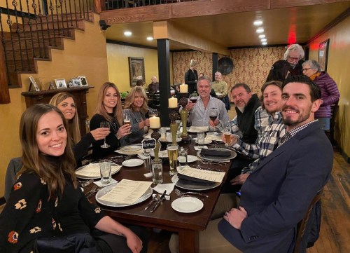 A group of eight people at a dinner table, smiling and toasting with drinks in a cozy restaurant setting.