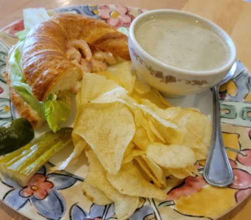 A plate with a shrimp croissant sandwich, potato chips, a pickle, and a bowl of creamy soup.