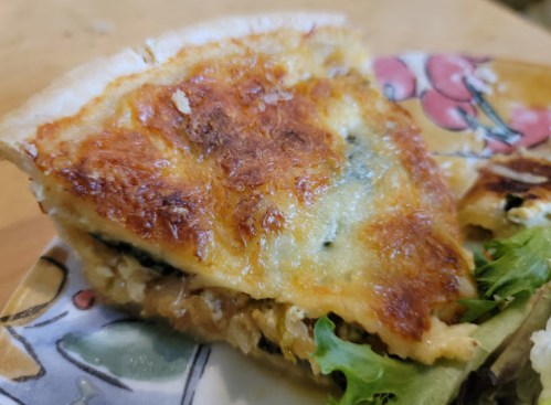 A slice of golden-brown quiche on a decorative plate, with a side of fresh greens.