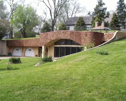 A unique, modern house with a curved roof, large windows, and a grassy lawn, surrounded by trees.