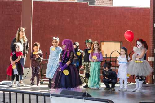 A group of children in costumes stands on stage, with a woman supervising and a red balloon in the background.