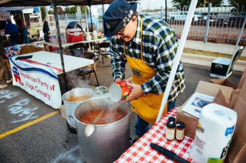 A man in an apron stirs a large pot of chili at an outdoor event, with tables and tents in the background.