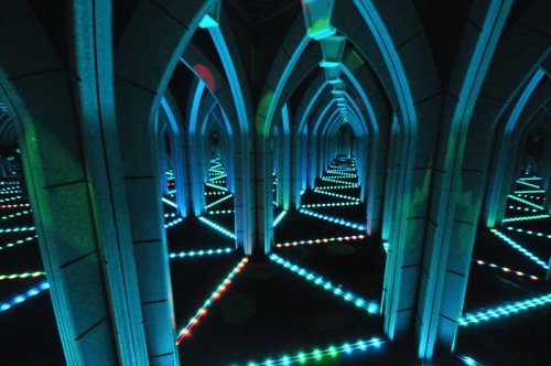 A mirrored corridor with colorful lights creating a geometric pattern, featuring archways and reflections.