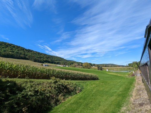 A scenic landscape featuring green fields, corn rows, and rolling hills under a bright blue sky with wispy clouds.
