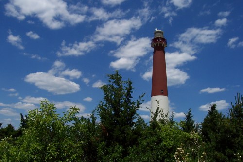 A tall red and white lighthouse stands among green trees under a blue sky with fluffy white clouds.