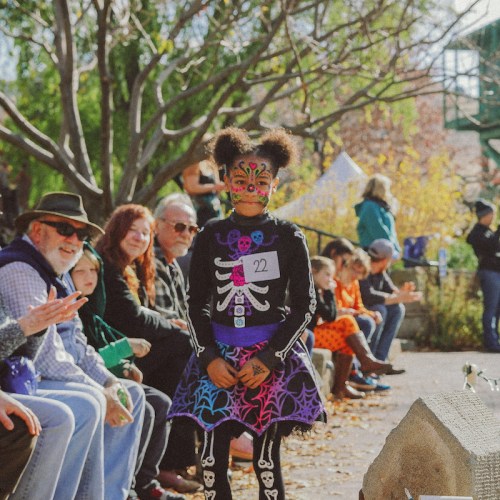 A child in a colorful skeleton costume sits among a crowd, surrounded by autumn foliage and festive decorations.