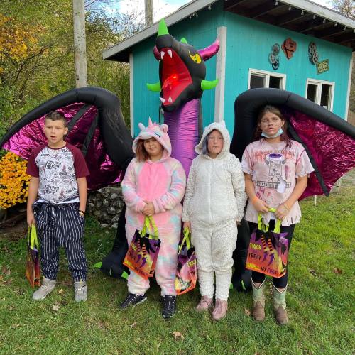 Four children in colorful costumes stand in front of a large inflatable dragon, holding treat bags.