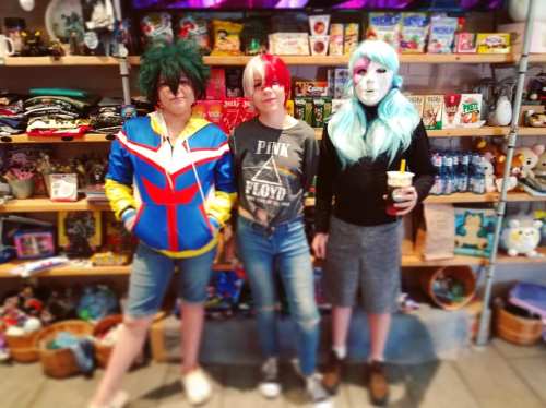 Three individuals in cosplay stand in a colorful shop, surrounded by merchandise and snacks. One wears a mask.
