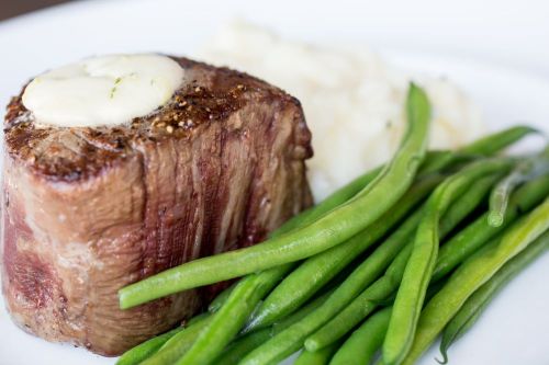 A perfectly cooked steak topped with butter, served with green beans and creamy mashed potatoes on a white plate.