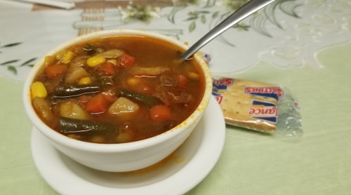 A bowl of hearty vegetable soup with a spoon, accompanied by a packet of crackers on a table.