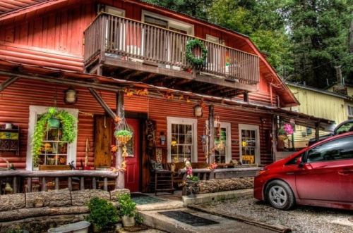 A charming red cabin with a balcony, adorned with greenery and flowers, surrounded by trees and a parked red car.