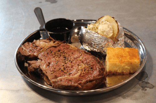 A plated meal featuring a juicy prime rib, baked potato with sour cream, and a piece of cornbread on a metal tray.