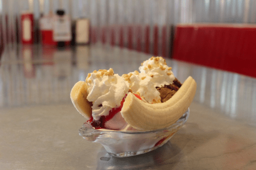 A banana split with scoops of chocolate and vanilla ice cream, topped with whipped cream and crushed nuts in a glass dish.
