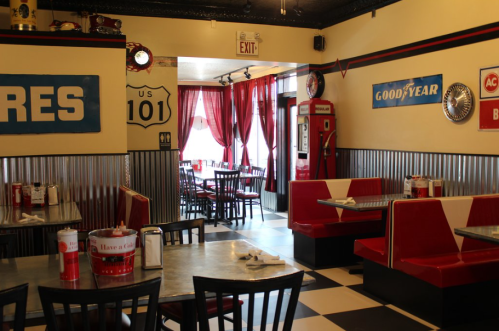 A retro diner interior with red booths, checkered floor, and vintage signs on the walls. Tables are set for dining.