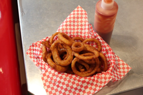 A basket of golden-brown onion rings on red and white checkered paper, next to a bottle of dipping sauce.