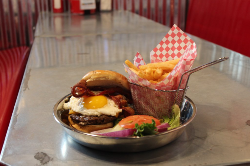 A burger topped with an egg and bacon, served with fries in a basket, on a metal table with red seating.