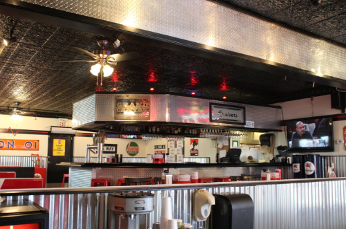 Interior of a diner featuring a bar area, red booths, and a TV displaying a show, with metallic decor and bright lighting.