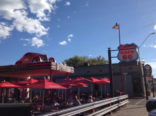 A retro diner with a car on the roof, outdoor seating, and a sign reading "Cruiser's Cafe 66" under a blue sky.
