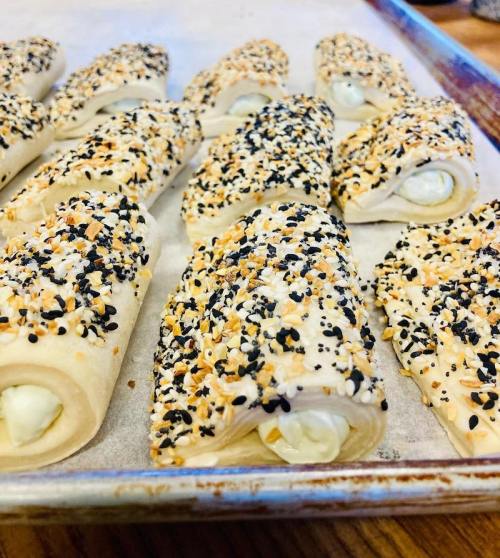 Baked pastry rolls topped with sesame seeds and filled with cream, arranged on a baking tray.