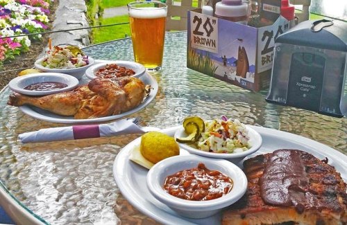 A table set with BBQ chicken, ribs, sides of coleslaw and beans, a lemon, and a beer in a scenic outdoor setting.