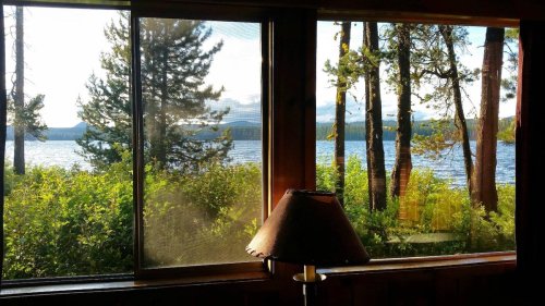 View of a serene lake surrounded by trees, seen through a window with a lamp in the foreground.