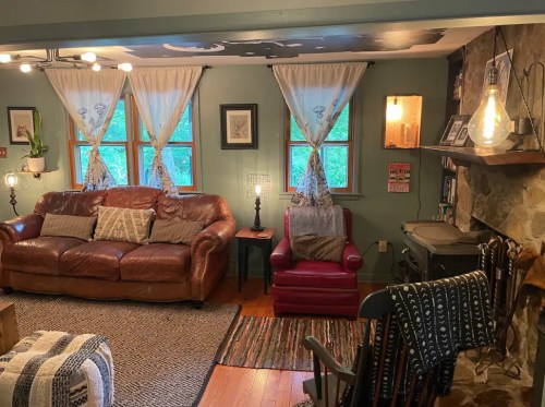 Cozy living room with a leather sofa, red armchair, stone fireplace, and large windows adorned with sheer curtains.