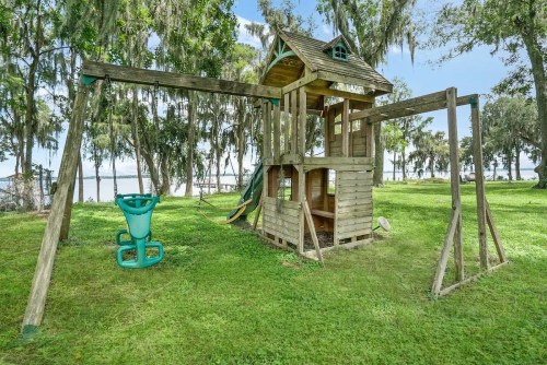 A wooden playset with a slide and swings, set on green grass near a lake and surrounded by trees.