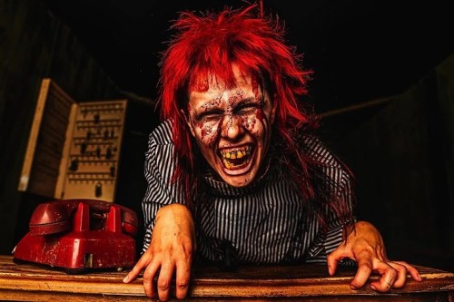 A person with wild red hair and a gruesome expression leans over a table, surrounded by eerie decor.