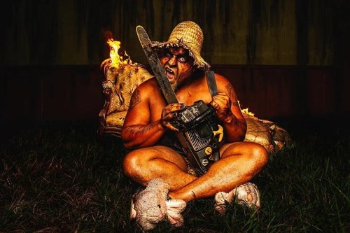 A muscular figure in a straw hat sits on a burning chair, holding a chainsaw, surrounded by grass and flames.