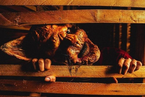 A creepy, grotesque mask peeks through wooden slats, with hands gripping the edges, creating a chilling atmosphere.