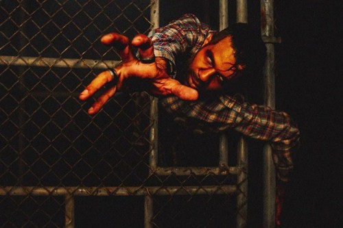 A man with a distressed expression reaches through a chain-link fence, his hand bloodied and grasping.