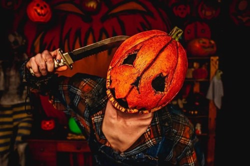 A person with a carved pumpkin for a head holds a knife, set against a backdrop of Halloween decorations.