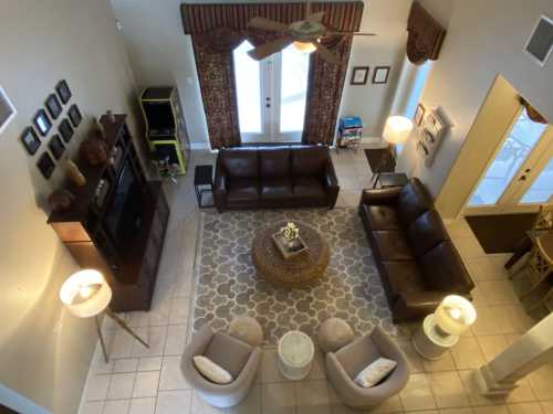 A spacious living room with brown sofas, a round coffee table, and large windows, viewed from above.