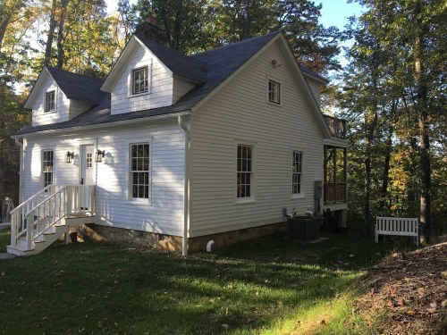 A white two-story house surrounded by trees, featuring a porch and a small bench in the yard.