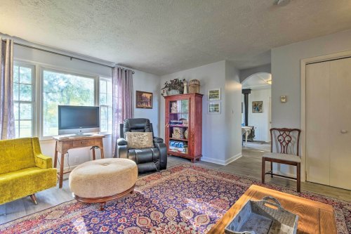Cozy living room with a yellow chair, black recliner, TV, and colorful rug, featuring natural light from large windows.