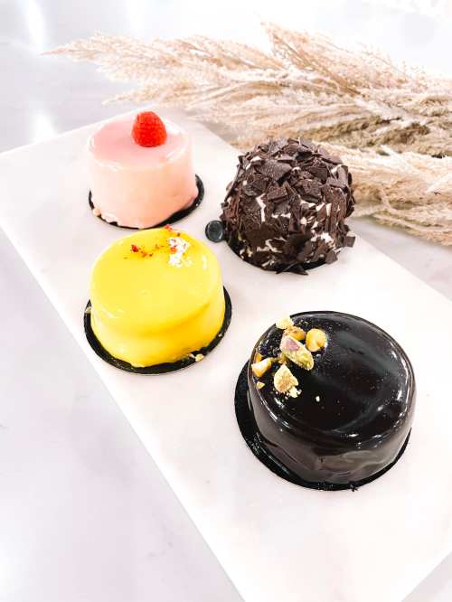 Four colorful desserts on a white platter: pink, yellow, black, and chocolate, with decorative dried grass in the background.