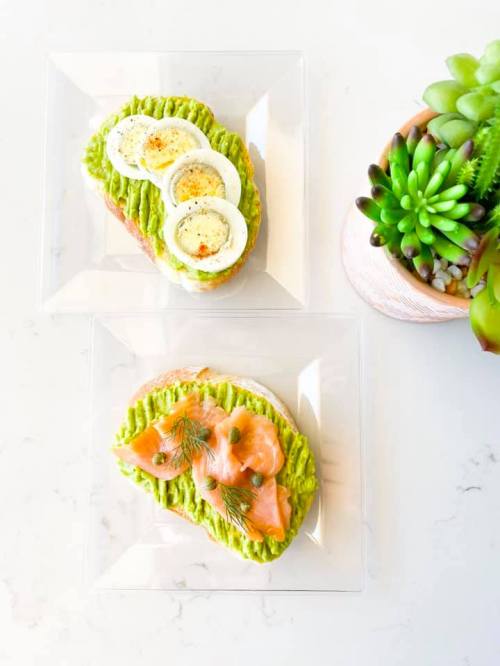 Two plates of avocado toast: one topped with sliced boiled eggs, the other with smoked salmon and dill.