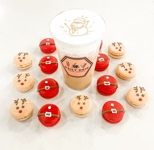 A festive arrangement of red and beige macarons around a cup of coffee with a snowman design on the foam.