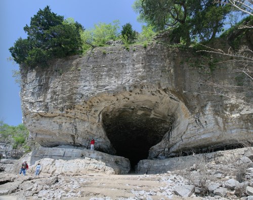 A large rock formation with a cave entrance, surrounded by greenery and people exploring the area.