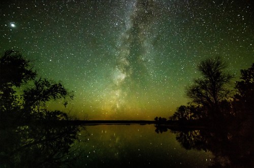 A serene night sky filled with stars and the Milky Way, reflecting on a calm lake surrounded by trees.