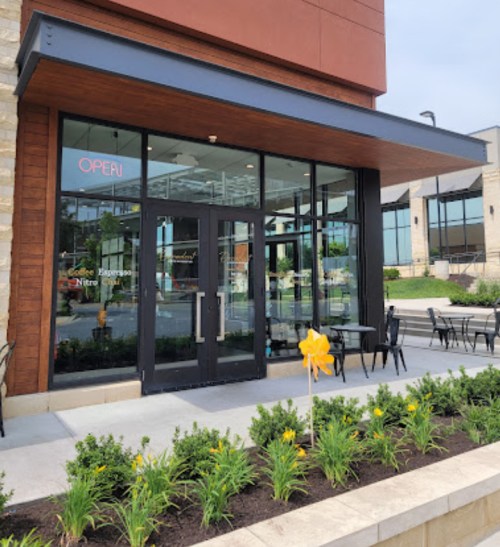 Exterior view of a modern café with large glass doors, outdoor seating, and a yellow ribbon decoration.