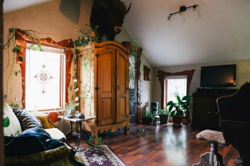 Cozy room with wooden furniture, plants, and a bison head mounted on the wall, featuring large windows and warm lighting.