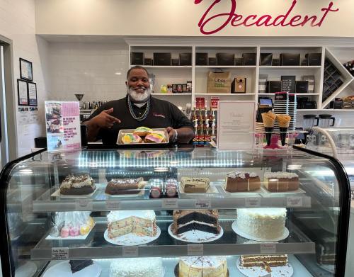 A smiling man stands behind a dessert display case filled with various cakes and pastries in a bakery.