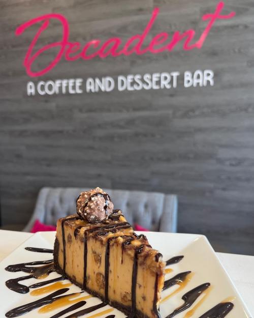 A slice of chocolate dessert topped with a truffle, served on a plate with chocolate sauce, against a café backdrop.