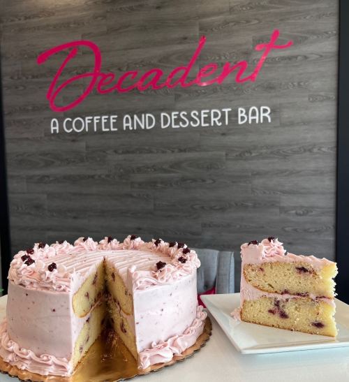 A pink frosted cake with a slice cut out, displayed in front of a "Decadent" coffee and dessert bar sign.